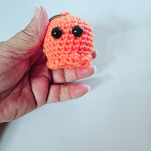 May include: A small, bright orange crocheted keychain shaped like a rounded character with two large, smooth, black plastic bead eyes. The textured yarn surface features a dense, uniform stitch pattern, and a metal ring is attached to the top.
