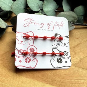 Puede incluir: Dos pulseras de hilo rojo con detalles anudados se exhiben en una tarjeta con el texto "String of fate" y "PERLEN.FACTORY". La tarjeta presenta ilustraciones de gatos de la suerte. El fondo es una superficie de madera.