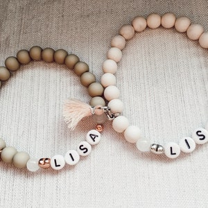 Puede incluir: Dos pulseras de cuentas con el nombre "Lisa" escrito en cuentas blancas. Una pulsera está hecha de cuentas de color marrón claro y la otra de cuentas blancas.