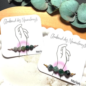 Puede incluir: Dos pulseras de cuentas con piedras verdes y grises se exhiben en tarjetas blancas con el texto "Armband des Neuanfangs". Las pulseras están sobre una bandeja blanca festoneada, con hojas de eucalipto en el fondo.