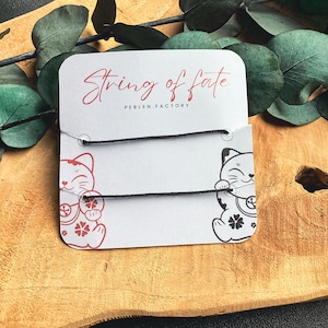 Puede incluir: Dos pulseras de cuerda negra con una tarjeta blanca que dice "String of Fate" y "Perlen.Factory" con una imagen de dibujos animados de un gato japonés de la suerte rojo y negro a cada lado.