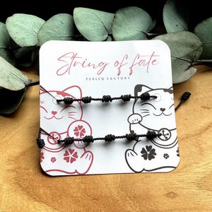 Puede incluir: Dos pulseras de cuerda negra con múltiples nudos se muestran en una tarjeta con el texto "String of fate" y "Perlen.factory". La tarjeta presenta ilustraciones de un gato de la suerte. El fondo incluye hojas verdes y una superficie de madera.