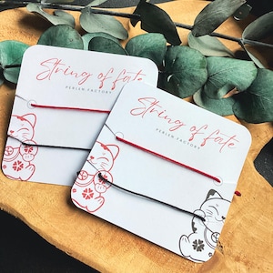 Könnte beinhalten: Zwei Karten mit roten und schwarzen Schnurarmbändern. Die Karten tragen den Text "String of fate" und eine Zeichnung einer Glückskatze. Der Hintergrund umfasst grüne Blätter und eine Holzoberfläche.