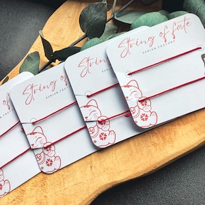 Könnte beinhalten: Vier rote Schicksalsbänder mit einem Glückskatzen-Anhänger an jedem. Die Armbänder befinden sich auf weißen Karten mit dem Text "String of fate" und "Perlen Factory".