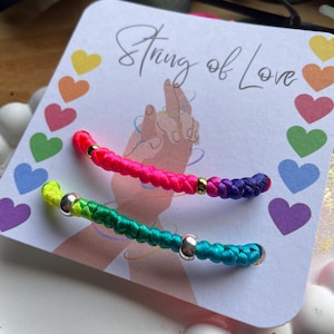 Puede incluir: Dos pulseras de la amistad coloridas con detalles plateados se muestran en una tarjeta que dice "String of Love". Las pulseras son en tonos rosa, morado, verde y azul, con cuentas doradas y plateadas.