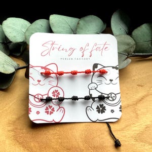 Puede incluir: Dos pulseras de cuerda anudada, una roja y otra negra, exhibidas en una tarjeta con una ilustración de gato de la suerte. La tarjeta dice "String of fate" y "PERLEN FACTORY". El fondo incluye hojas de eucalipto.