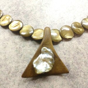 Puede incluir: Collar con cuentas doradas ovaladas y un colgante triangular de madera. El colgante tiene una perla blanca grande de forma irregular. El collar se muestra sobre un fondo neutro.