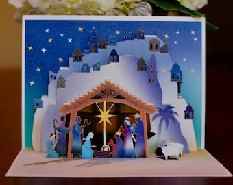 May include: A pop-up Christmas card depicting the Nativity scene. The card features a layered design with a starry night sky, buildings, and figures representing the Holy Family and the Three Wise Men. The color palette includes blues, golds, and whites.