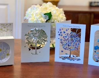 May include: Four sympathy cards with different designs. The cards feature a tree, flowers, and birds. The cards are white with silver and blue accents. The text on the cards reads "With Deepest Sympathy".
