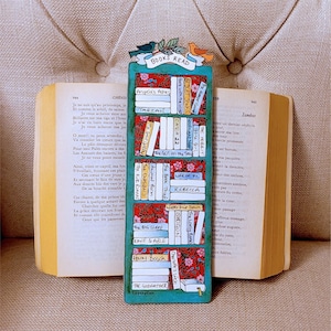 May include: A colorful bookmark shaped like a bookshelf, featuring miniature book titles like "Angela's Ashes" and "The Hobbit." The bookmark is teal with a floral pattern and topped with bird illustrations. It rests in an open book.