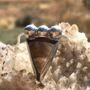 May include: A silver pendant with a triangular brown agate stone and three silver beads. The pendant is resting on a bed of white crystals.