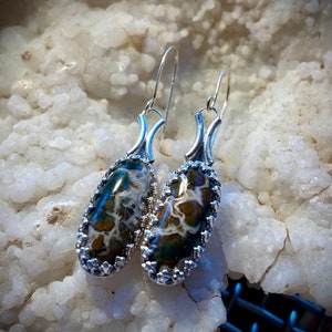 May include: A pair of silver dangle earrings with oval-shaped green, brown, and white agate stones set in a silver filigree design.