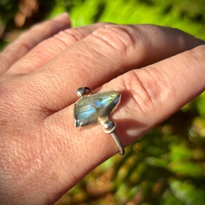 May include: A silver ring featuring a labradorite gemstone. The gemstone has a blue and green iridescent sheen. The ring is set on a finger, with a blurred green background.