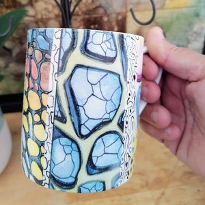 May include: A white ceramic mug with a colourful geometric pattern. The pattern features blue, yellow, pink and brown shapes.