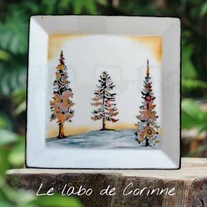 May include: Square white ceramic plate with a black border. The plate features a hand-painted design of three stylized pine trees in shades of orange, pink, and white against a yellow and blue background. The text "Le labo de Corinne" is at the bottom.