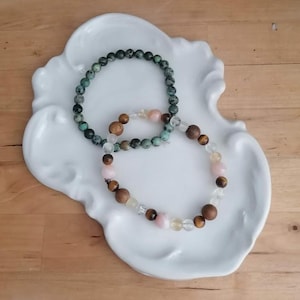 Op de afbeelding: Twee armbanden met kralen liggen op een wit, geschulpte schaal. Eén armband heeft donkergroene kralen, de andere combineert roze, bruine, heldere en goudkleurige kralen. De armbanden worden gepresenteerd op een licht houten oppervlak.