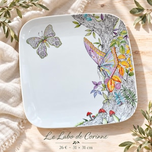 May include: A square white ceramic plate featuring a detailed illustration of a tree trunk with colourful butterflies, green leaves, and small red mushrooms. The plate includes the text Le Labo de Corinne and dimensions of 31 x 31 cm.