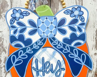 May include: A wooden pumpkin door hanger with a blue and white bow, floral details, and the word "Hey" in a blue and white plaid pattern.