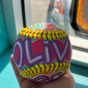 Può includere: Una palla da softball colorata con il nome "OLIV" in grandi lettere stilizzate. La palla presenta un design vibrante con accenti rosa, viola, gialli e turchesi. Le cuciture sono gialle e l'estetica generale è giocosa e personalizzata.