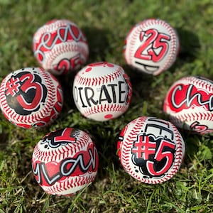 May include: Six white baseballs with red stitching, each featuring hand-painted designs in red, black, and white. The balls display various custom graphics, including the text PIRATE, #3, #5, and #25, along with stylised lettering and numbers.
