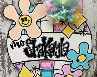 Può includere: Un appendiporta rotondo bianco con un motivo geometrico colorato. L'appendiporta presenta un grande testo nero che dice "Mrs. Shakaya" e diversi fiori colorati con facce sorridenti. Un grande fiocco rosa e verde è attaccato alla parte superiore dell'appendiporta.
