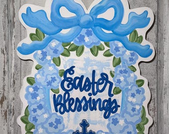 May include: A white wooden sign with a blue ribbon bow and a wreath of blue flowers. The sign says "Easter Blessings" and has a blue cross in the center.
