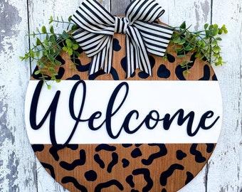 May include: A round wooden door hanger with a black and white leopard print design. The door hanger has a white stripe with the word "Welcome" in black lettering. A black and white striped bow is attached to the top of the door hanger.