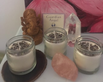 Puede incluir: Tres pequeños frascos de vidrio con velas blancas y flores de lavanda secas en la parte superior. Las velas están sobre un posavasos de madera. Hay un cristal de cuarzo rosa y un cristal transparente junto a las velas. Una pequeña estatua de madera de Ganesha está en el fondo. La tarjeta en el fondo dice "Ángel de la Guarda".