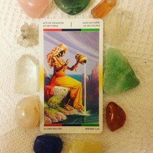 Puede incluir: Carta de tarot que representa el As de Copas, con una mujer vertiendo agua de una jarra en una piscina de agua. La carta est&aacute; rodeada de varios cristales, incluyendo cuarzo claro, cuarzo rosa y aventurina verde.