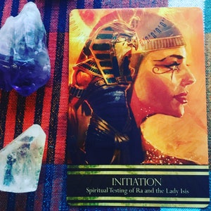 Puede incluir: Una carta de tarot con un fondo dorado y una mujer con un tocado dorado. La carta se titula "Iniciaci&oacute;n: Prueba Espiritual de Ra y la Dama Isis".