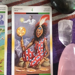Puede incluir: Carta de tarot que representa el Cuatro de Pent&aacute;culos. La carta presenta una figura sentada que lleva una t&uacute;nica con estampado rojo y blanco y un tocado con plumas. La figura sostiene una moneda de oro con un dise&ntilde;o de flor. La carta tambi&eacute;n presenta una figura blanca en el fondo que sostiene una l&aacute;mpara encendida.