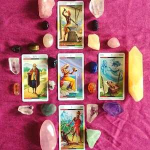 Puede incluir: Un despliegue de cartas de tarot con varios cristales sobre un fondo de tela rosa. Las cartas son de la baraja "Tarot of the Divine" e incluyen las cartas "As de Espadas", "XIV Templanza", "I Copas", "II Espadas" y "III Varitas".