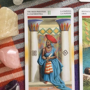 Puede incluir: La carta del tarot La Sacerdotisa muestra a una mujer con un sombrero rojo y una t&uacute;nica azul, sentada en un trono con dos pilares detr&aacute;s de ella. La carta es del mazo "The Sacred Rose Tarot" e incluye el texto "The High Priestess", "La Sacerdotisa", "II", "La Papessa", "La Papesse", "Die Hohepriesterin", "De Hogepriesteres", y "Pentacli Oros".