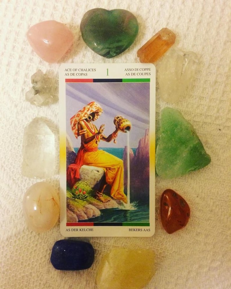 Puede incluir: Carta de tarot que representa el As de Copas, con una mujer vertiendo agua de una jarra en una piscina de agua. La carta est&aacute; rodeada de varios cristales, incluyendo cuarzo claro, cuarzo rosa y aventurina verde.
