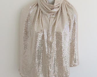 May include: A champagne-coloured scarf with a cowl neck design. The scarf is covered in small, reflective sequins, creating a textured, sparkling effect. The fabric is draped over a mannequin, showcasing its shape and drape.