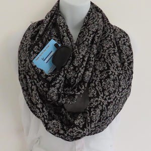 Puede incluir: Bufanda infinita con estampado en blanco y negro con un bolsillo oculto. La bufanda está drapeada sobre un maniquí y presenta un diseño floral. Un pequeño objeto negro y una tarjeta azul son visibles en el bolsillo.