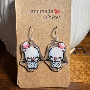 May include: A pair of handmade earrings featuring a cartoon character with a white body, pink accents, and black wings. The earrings are attached to a brown card with the text "Handmade with love".