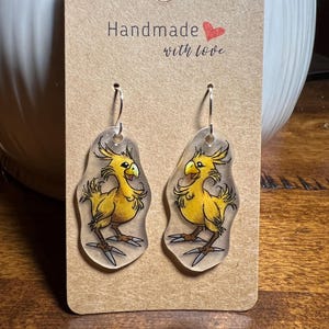 May include: A pair of handmade earrings featuring yellow cartoon birds with black and gray details. The earrings are on a brown card with the text "Handmade with love". The earrings have silver hooks.