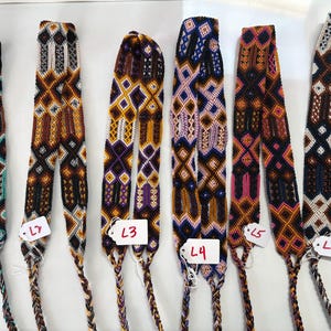 May include: Six long, woven fabric straps featuring intricate geometric patterns in various color combinations including teal, orange, purple, pink, and black. Each strap is labeled with a small white tag marked L1 through L6 and ends in a braided cord.
