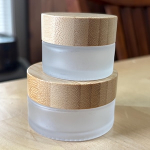 May include: Two cylindrical cosmetic jars with frosted glass bases and light brown bamboo lids. The jars are stacked vertically, with the smaller jar resting on top of the larger jar. Both containers feature a smooth, matte finish on the glass.