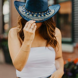 May include: A woman wearing a blue denim cowboy hat with beaded trim, a white tank top, a brown belt with double O-rings, and blue denim jeans.