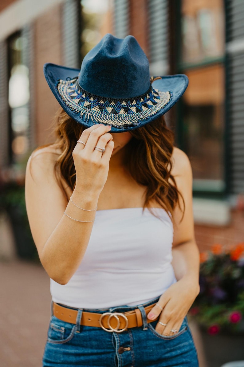 May include: A woman wearing a blue denim cowboy hat with beaded trim, a white tank top, a brown belt with double O-rings, and blue denim jeans.