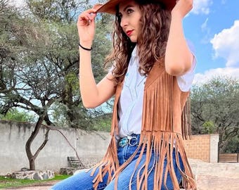 May include: A brown suede cowboy hat and fringed vest are worn with a white shirt and blue jeans. The vest has long fringe. The jeans have ripped knees. A turquoise belt buckle adds a pop of color. The outfit is a western-inspired style.