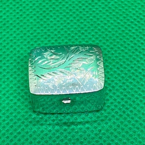 Könnte beinhalten: Kleine, quadratische, silberfarbene Schatulle mit Scharnierdeckel. Der Deckel ist mit einem gravierten Blumenmuster verziert. Die Schatulle befindet sich vor einem grünen Hintergrund.