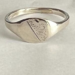 May include: A silver signet ring featuring a rounded band and a shield-shaped face. The face is split diagonally, with a textured section and a smooth section. The ring is likely made of sterling silver.