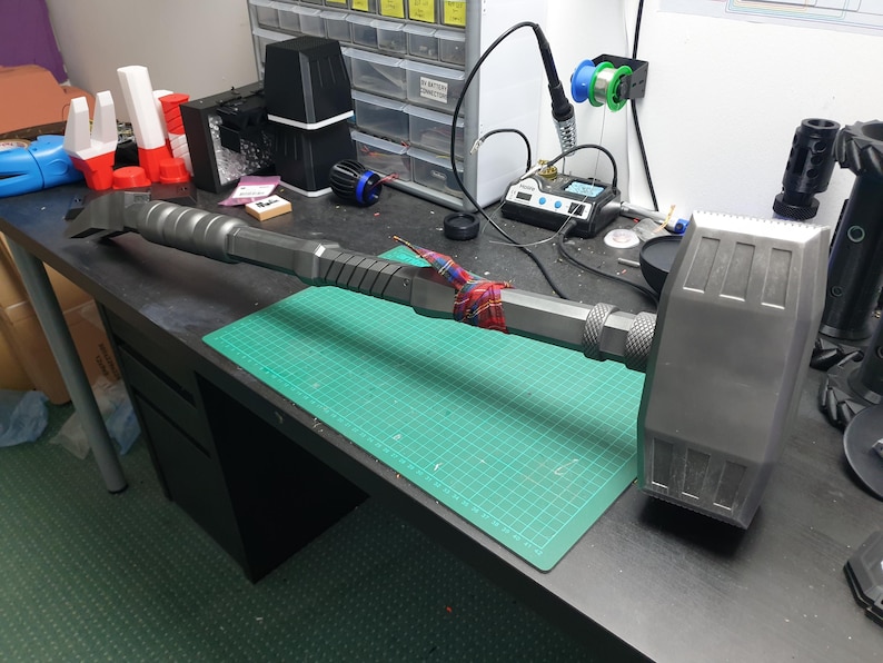 May include: A large, metallic grey prop hammer with a rectangular head and a cylindrical handle. The handle is wrapped with a red, blue, and yellow plaid fabric. The hammer is on a green cutting mat on a black desk.