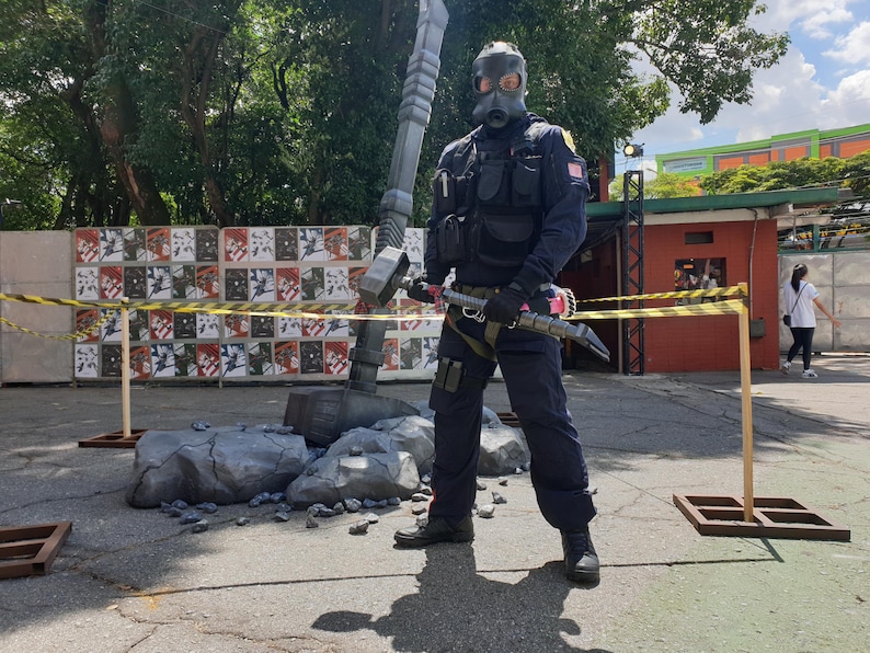 May include: A person wearing a black tactical vest and a gas mask is holding a large silver hammer. The hammer is resting on a pile of broken rocks. The person is standing in front of a wall with posters on it.