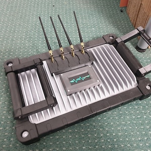 May include: A gray and black device with a handle and multiple antennas. The device has a small screen displaying a green waveform.
