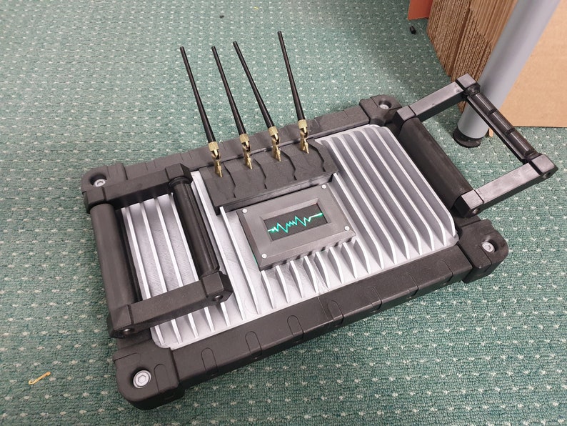 May include: A gray and black device with a handle and multiple antennas. The device has a small screen displaying a green waveform.