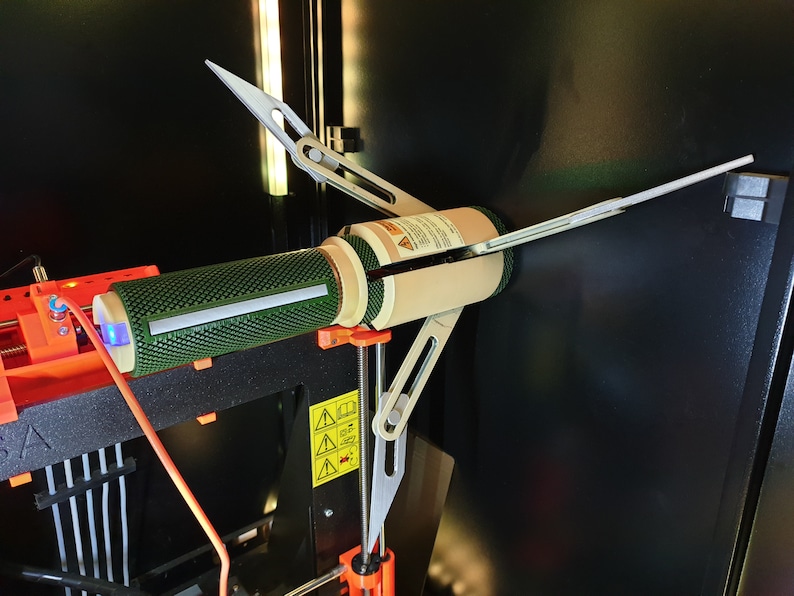 May include: A 3D printer component with a green mesh cylinder, beige cylindrical sections, and silver support arms. The device is mounted on an orange and black 3D printer frame, with a blue light visible.